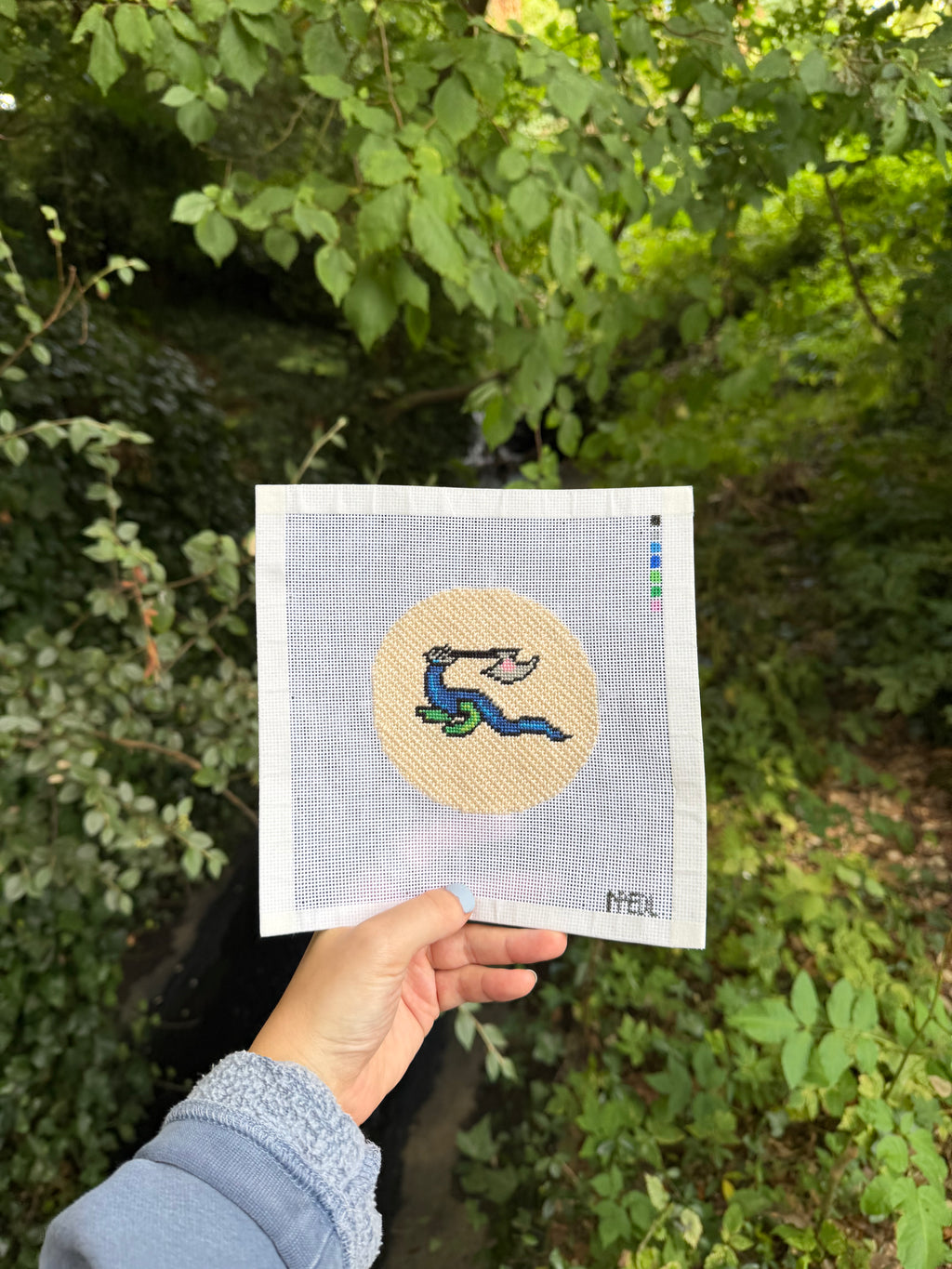 A hand holds a square needlepoint canvas featuring a circular beige design with a blue and green serpent motif, set against a lush green outdoor background.
