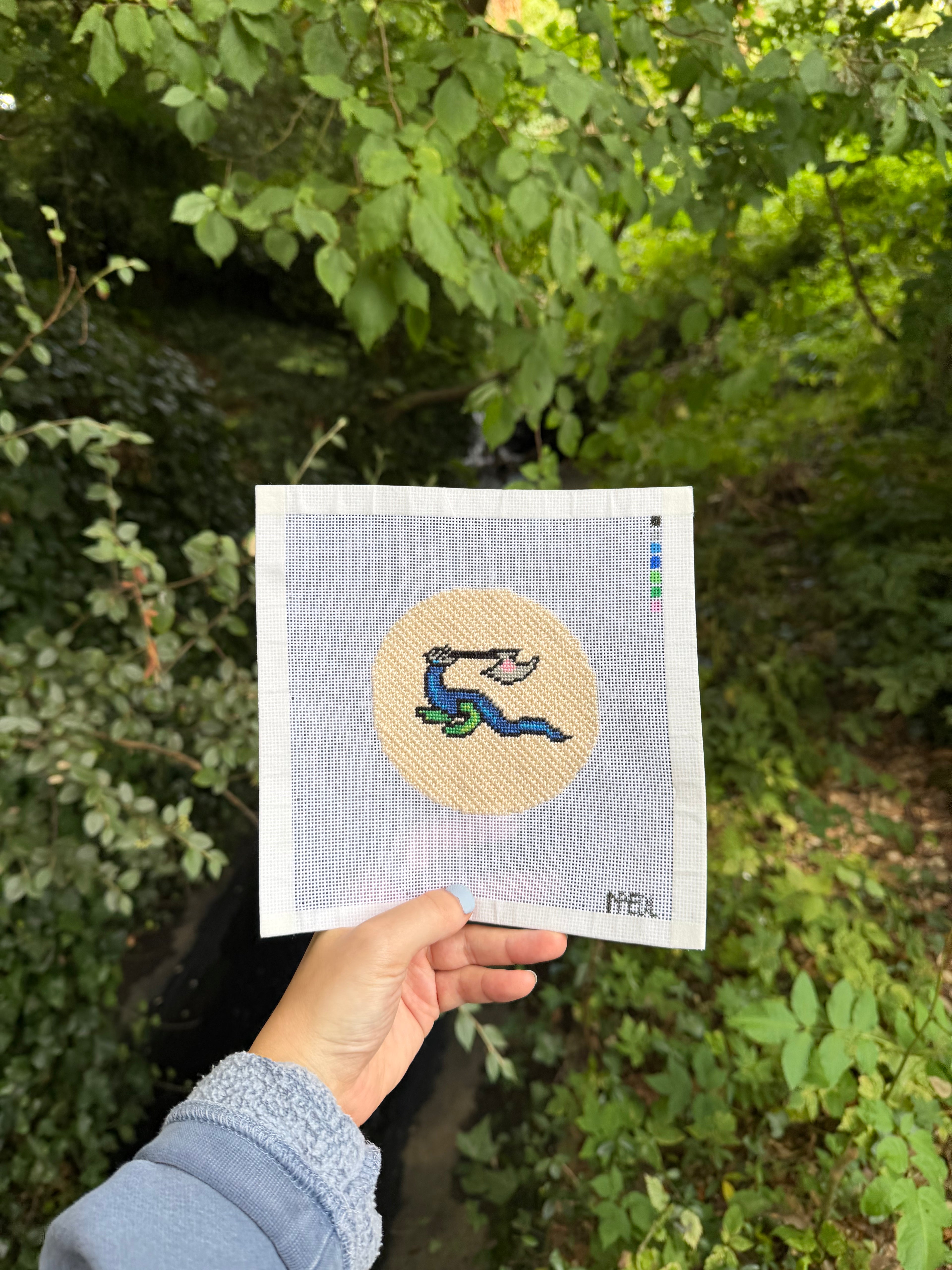 A hand holds a square needlepoint canvas featuring a circular beige design with a blue and green serpent motif, set against a lush green outdoor background.